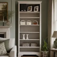 Tall 3ft handmade bookcase painted in off-white Purbeck Stone, standing against a sage green panelled wall beside a fireplace, its six shelves styled with framed prints, round mirrors, candlesticks and wicker baskets