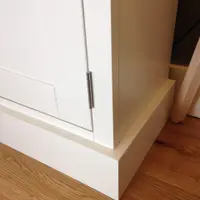 Close view of the lower corner of the white painted sideboard, showing the stepped plinth, crisp Shaker door frame and a brushed steel hinge against an oak floor