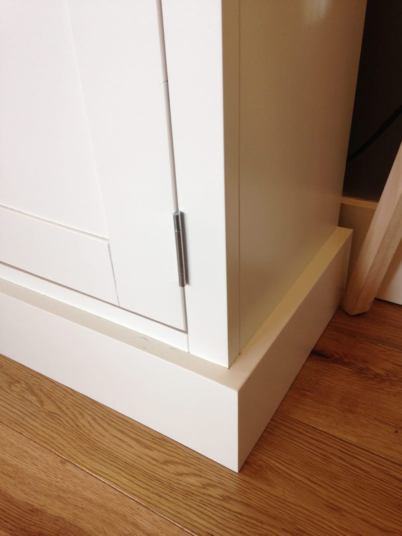 Close view of the lower corner of the white painted sideboard, showing the stepped plinth, crisp Shaker door frame and a brushed steel hinge against an oak floor