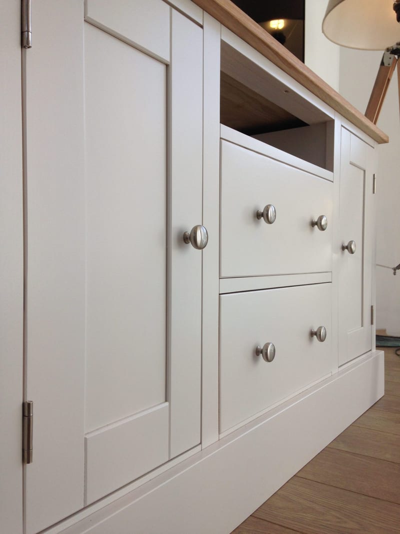 Low angled view along the front of the painted TV cabinet, showing the Shaker doors, central drawer stack, open media shelf and the overhang of the solid oak worktop