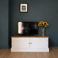 The 4ft Radwell TV base unit painted in cream with a solid oak worktop and two Shaker doors, standing against a dark teal wall with a flatscreen television and a glass vase of sunflowers on top