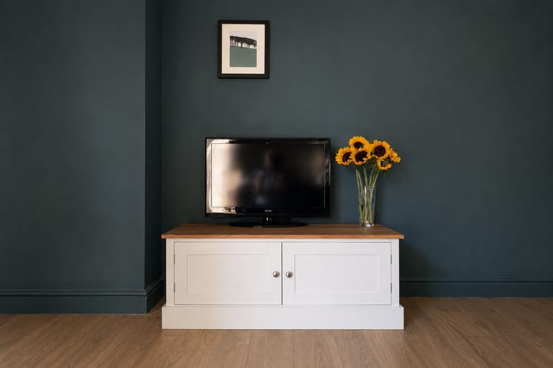 The 4ft Radwell TV base unit painted in cream with a solid oak worktop and two Shaker doors, standing against a dark teal wall with a flatscreen television and a glass vase of sunflowers on top