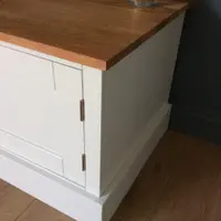 Close view of the corner of the TV stand, showing the sawn edge and overhang of the solid oak worktop above the painted carcass, with a brushed steel hinge and stepped plinth on an oak floor
