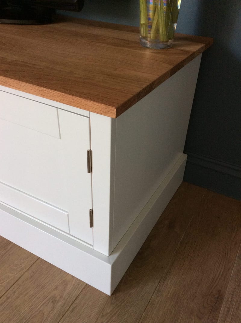 Close view of the corner of the TV stand, showing the sawn edge and overhang of the solid oak worktop above the painted carcass, with a brushed steel hinge and stepped plinth on an oak floor