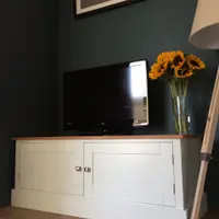 Angled view of the Radwell TV base unit in a dark teal room, both Shaker doors closed with brushed nickel knobs, below a television, sunflowers and a framed print of trees