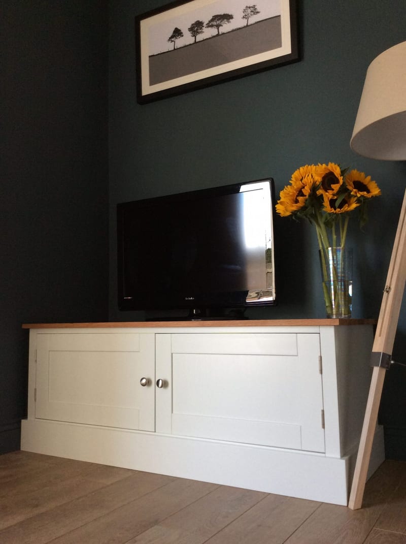 Angled view of the Radwell TV base unit in a dark teal room, both Shaker doors closed with brushed nickel knobs, below a television, sunflowers and a framed print of trees