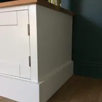 Side and corner view of the painted TV unit showing the plain painted end panel, the overhanging oak worktop, brushed steel hinges and the deep stepped plinth