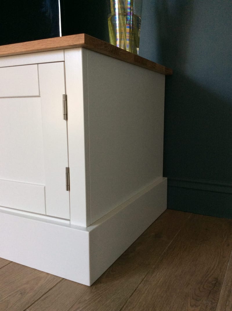 Side and corner view of the painted TV unit showing the plain painted end panel, the overhanging oak worktop, brushed steel hinges and the deep stepped plinth