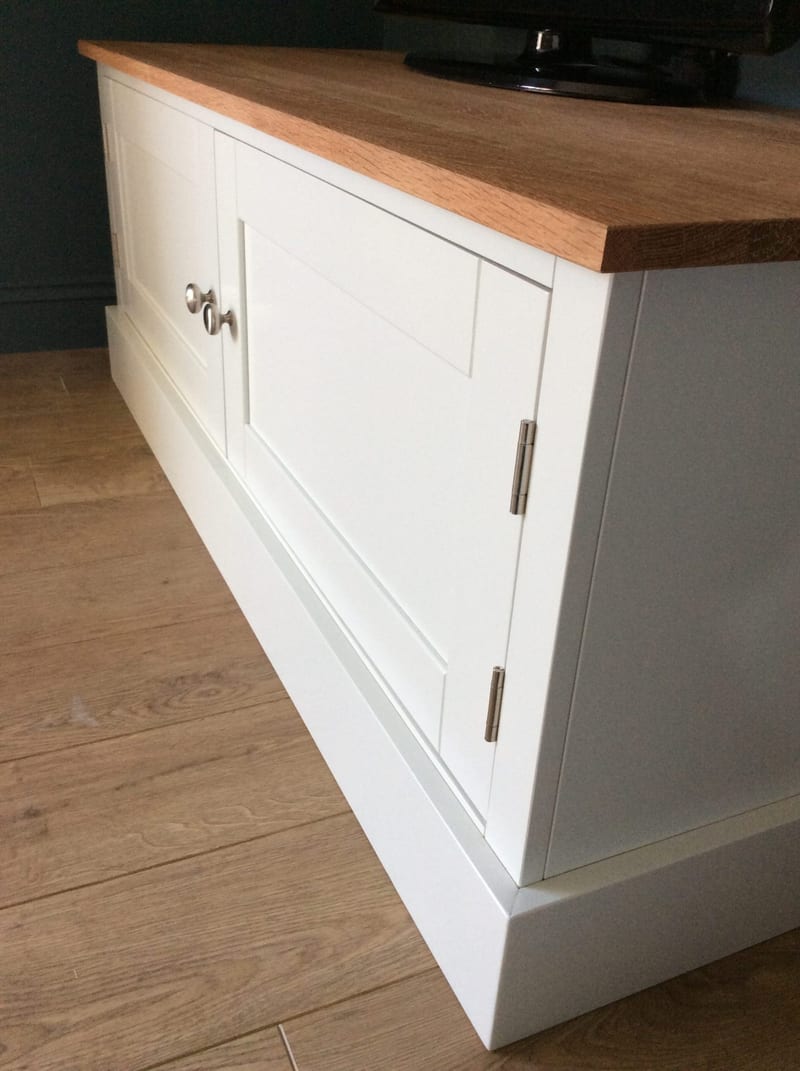 Angled corner view of the TV unit showing the solid oak worktop, one Shaker door with brushed nickel knob and hinges, and the painted plinth below