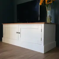 Three-quarter view of the 4ft Radwell TV base unit with both Shaker doors closed, a television and a vase of sunflowers on the oak worktop, against a dark teal wall
