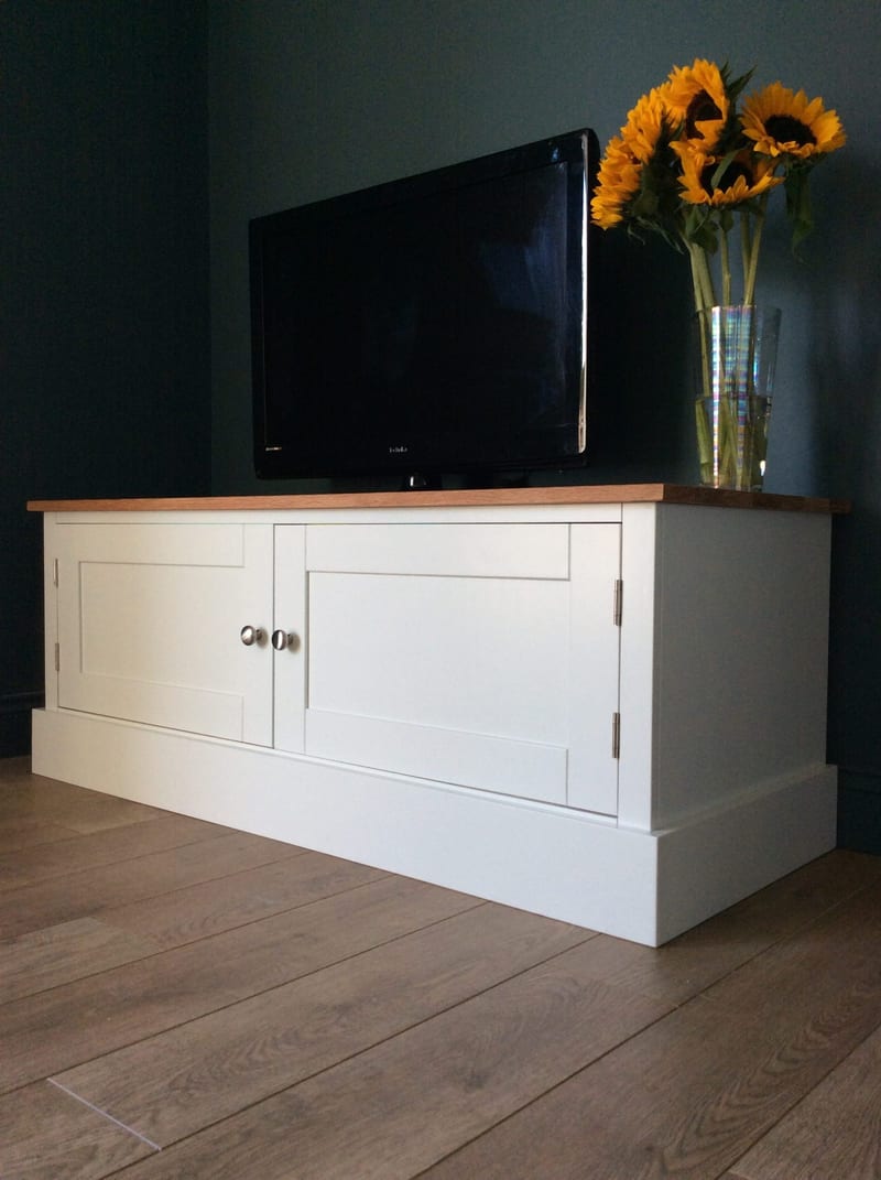 Three-quarter view of the 4ft Radwell TV base unit with both Shaker doors closed, a television and a vase of sunflowers on the oak worktop, against a dark teal wall