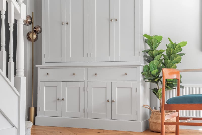 The grey painted Radwell dresser standing beside a white staircase, showing two wide drawers above four Shaker cupboard doors on a stepped plinth, with the upper doors continuing past the top of the frame, a fiddle leaf fig and a wooden chair alongside on a herringbone floor