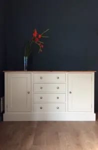 Full front view of the cream painted sideboard against a dark navy wall, two shaker doors flanking four drawers with brushed nickel knobs and a slim blue vase of red gladioli on the pine top