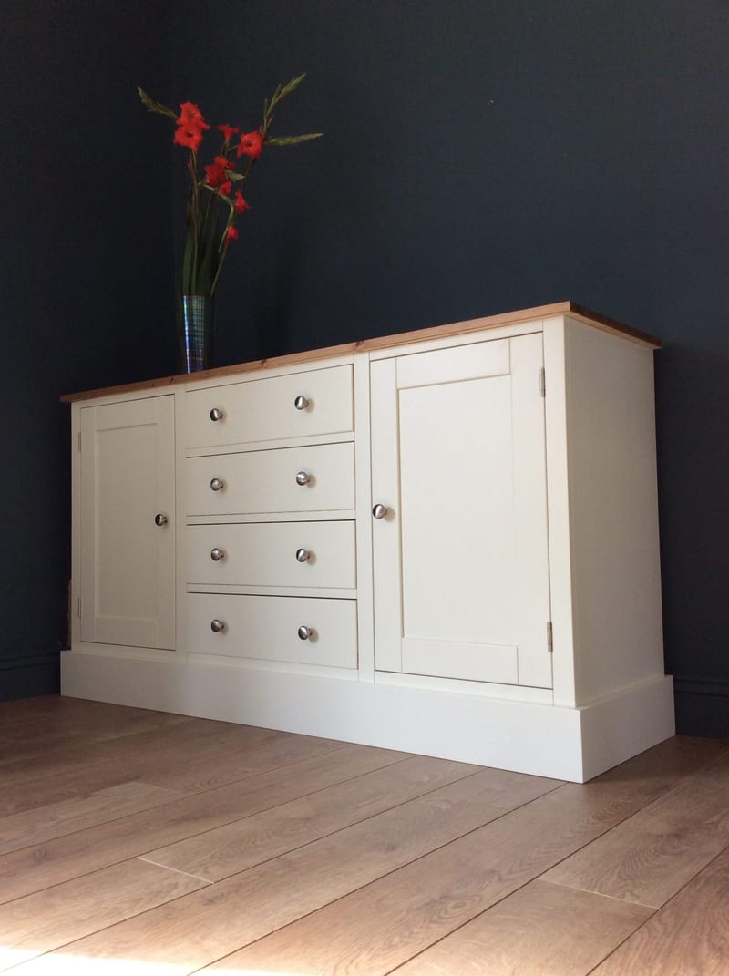 Angled view of the cream sideboard on oak laminate flooring, showing the honey pine top, four drawers with brushed nickel knobs between two shaker doors and the deep plinth base