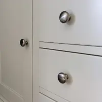 Close-up of two brushed nickel mushroom knobs on the cream drawer fronts beside the framed cupboard door, showing the fine shadow gaps between drawers and the smooth painted finish