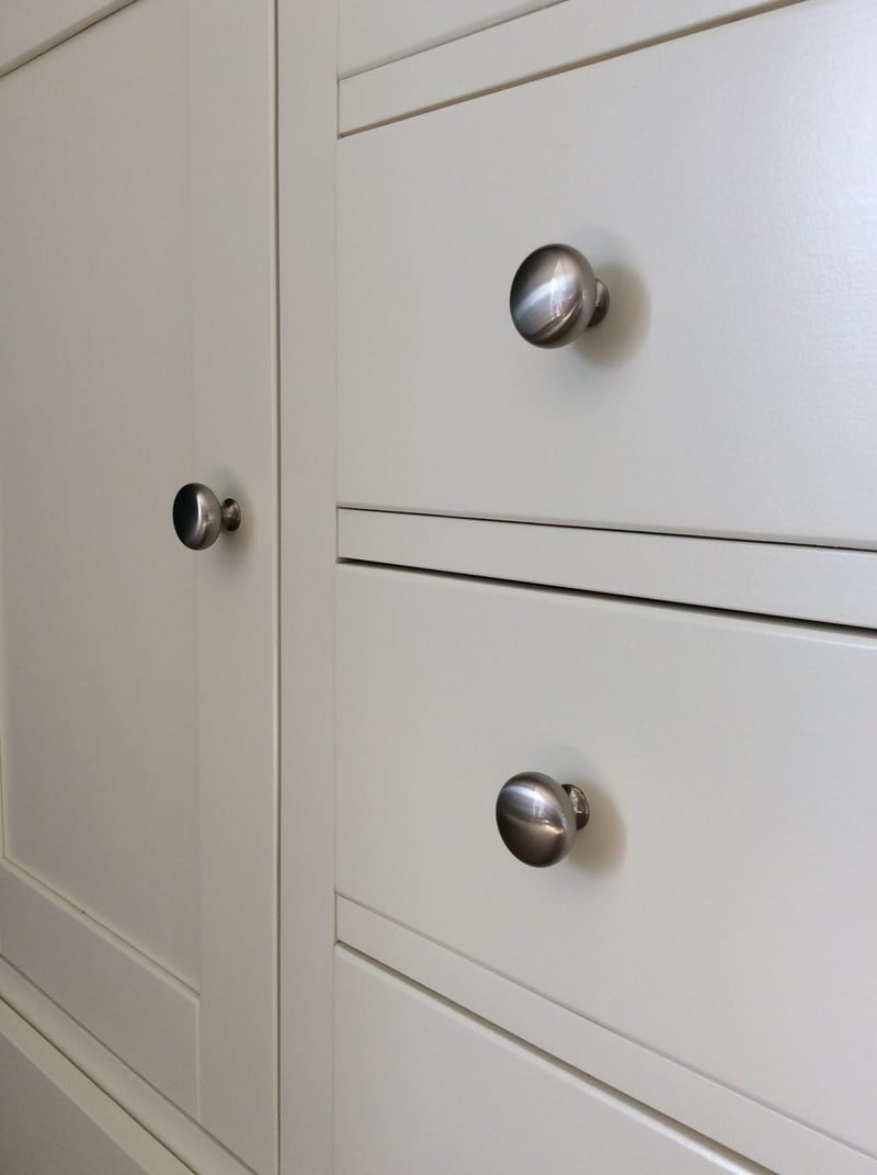 Close-up of two brushed nickel mushroom knobs on the cream drawer fronts beside the framed cupboard door, showing the fine shadow gaps between drawers and the smooth painted finish