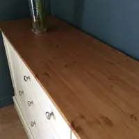 Looking down the length of the stained pine top, showing the knotty grain and board joins, a tall glass vase of red gladioli and the cream drawer fronts below against a blue-grey wall