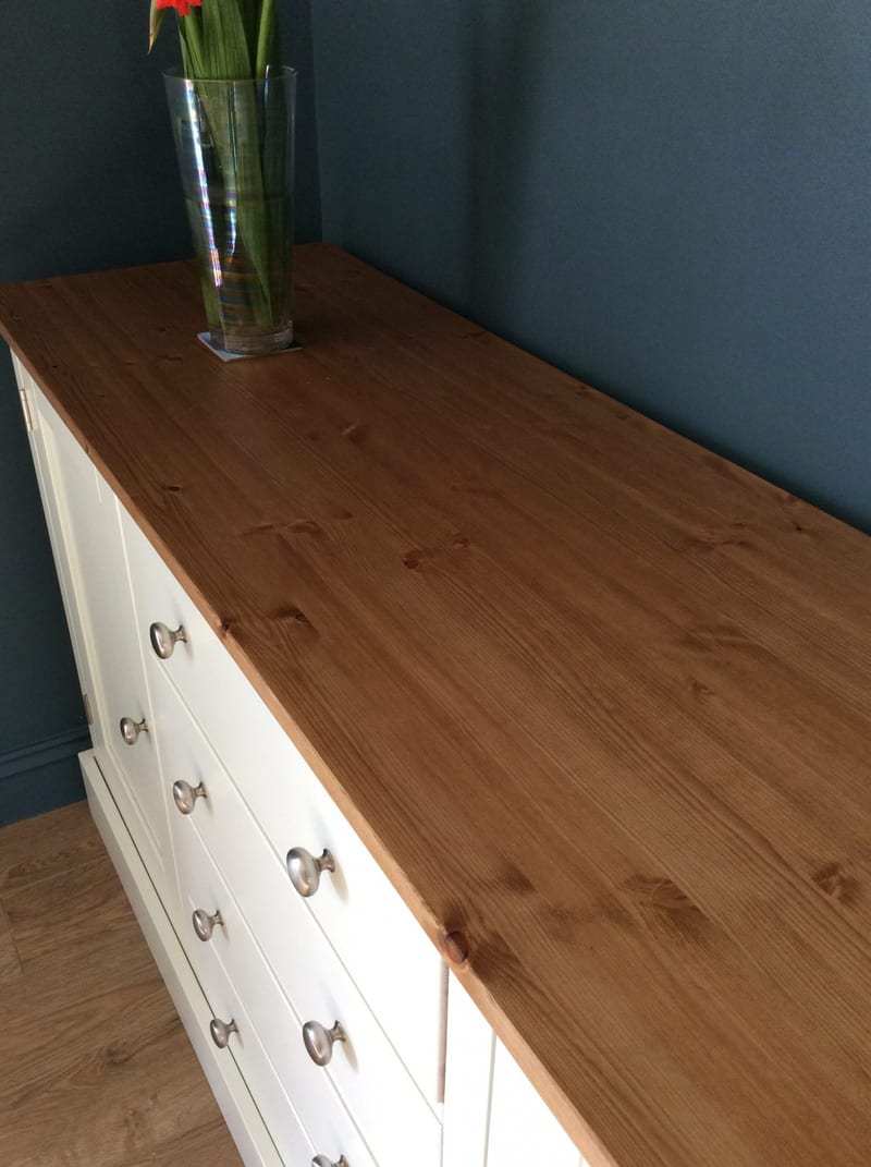 Looking down the length of the stained pine top, showing the knotty grain and board joins, a tall glass vase of red gladioli and the cream drawer fronts below against a blue-grey wall