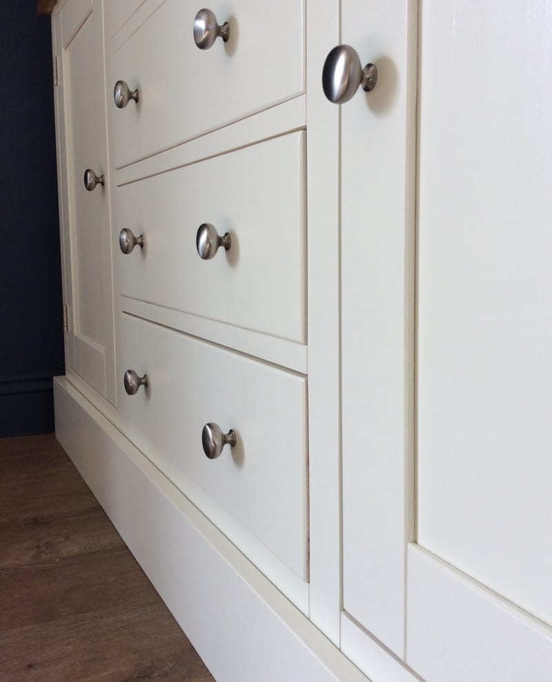 Sharp angled view along the cream painted front, showing three drawer fronts with pairs of brushed nickel knobs, a cupboard door knob in the foreground and the deep plinth running along the base