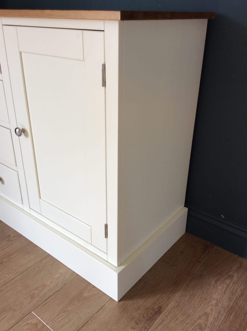 Corner view of the right-hand end of the cream sideboard, showing the plain side panel, the overhanging wooden top, a shaker door on brushed nickel hinges and the plinth on oak laminate flooring