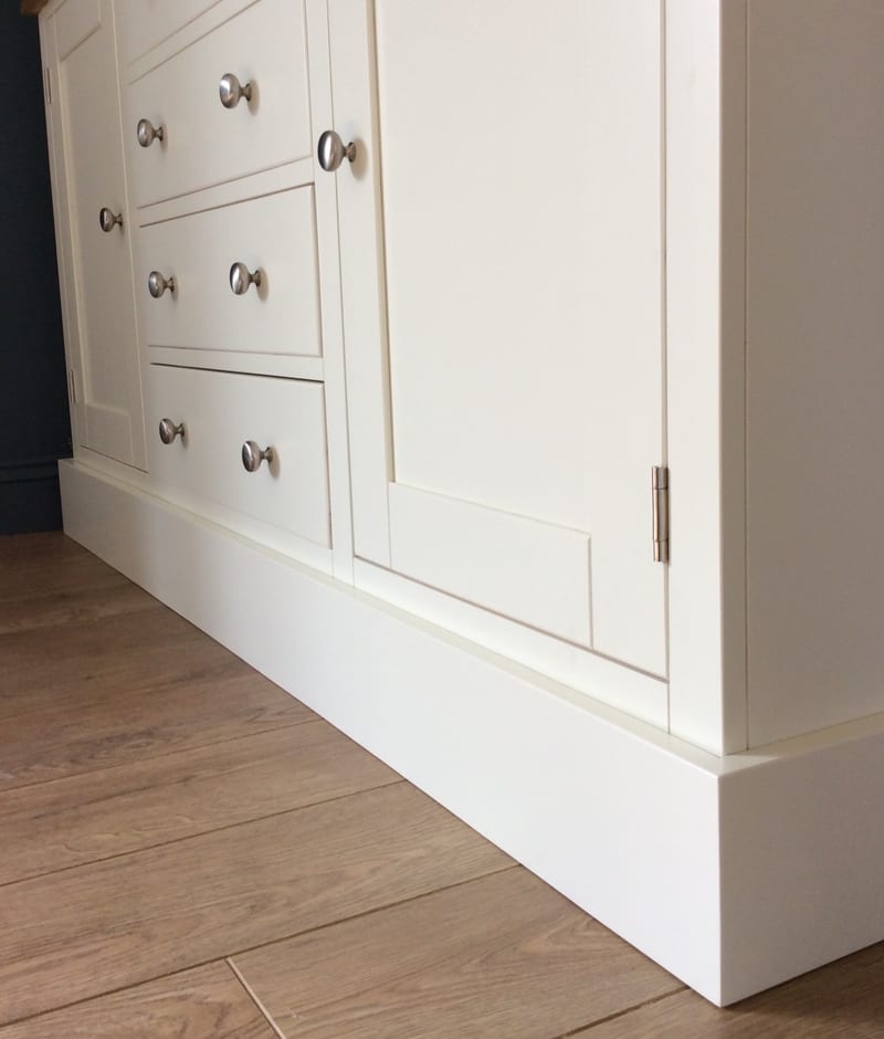 Low view of the base of the cream sideboard, showing the deep stepped plinth, the framed cupboard doors on brushed nickel hinges and the lowest drawers with their brushed nickel knobs
