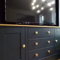 Close view of the navy base cupboard, showing three drawers and two shaker doors with aged brass knobs, the oak worktop edge and a television reflecting a starburst ceiling light