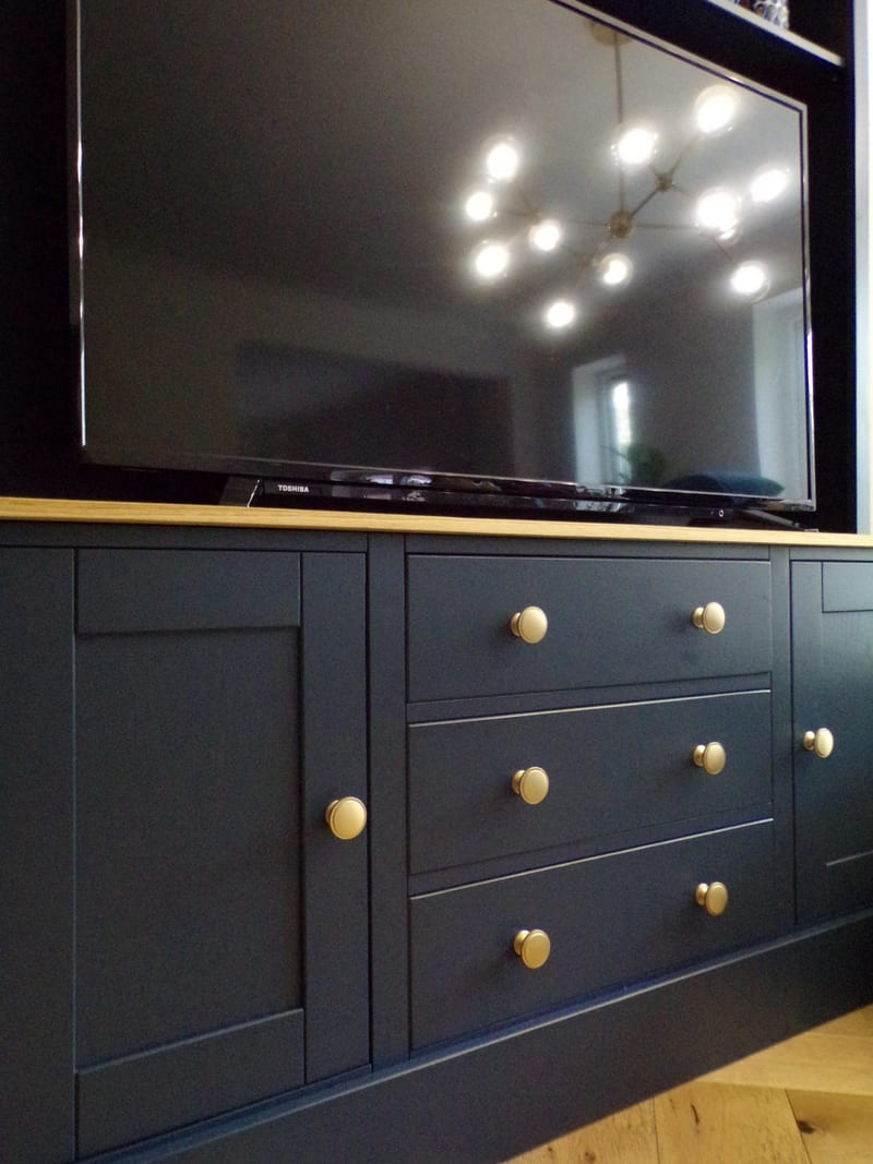 Close view of the navy base cupboard, showing three drawers and two shaker doors with aged brass knobs, the oak worktop edge and a television reflecting a starburst ceiling light