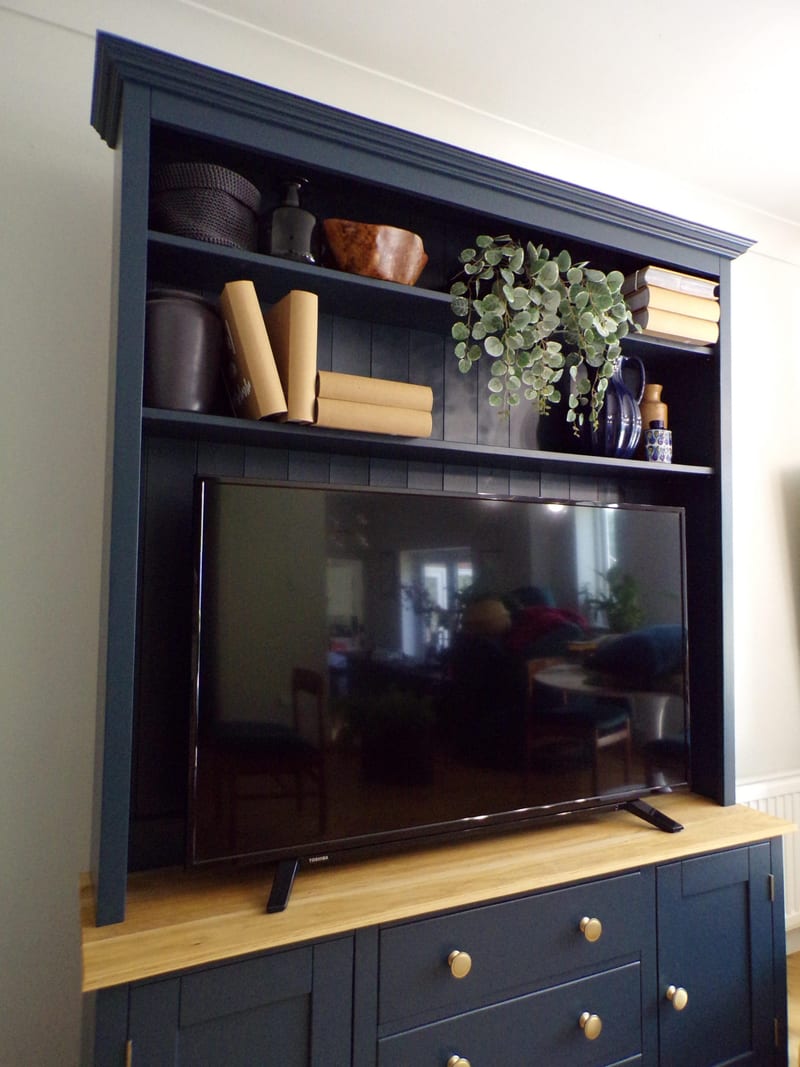 Angled view of the navy media unit showing the moulded cornice, two shelves styled with books, a trailing plant and pots, and the television standing on the oak worktop above brass knobs