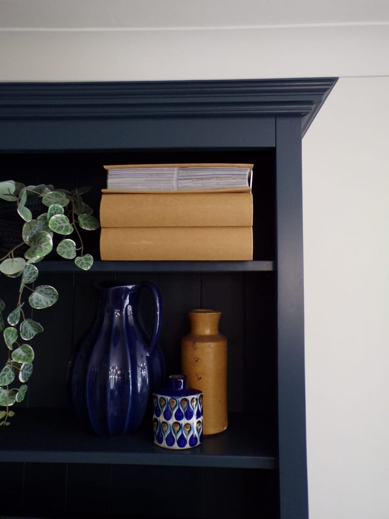 Detail of the top corner of the navy dresser rack, showing the layered cornice, tongue-and-groove backboards, stacked books, a cobalt jug, a mustard stoneware bottle and a patterned lidded pot