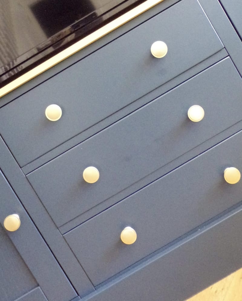 Overhead angled view across the navy drawer fronts, showing the round brass knobs, the crisp shadow gaps between the drawers and the lower edge of the television screen above