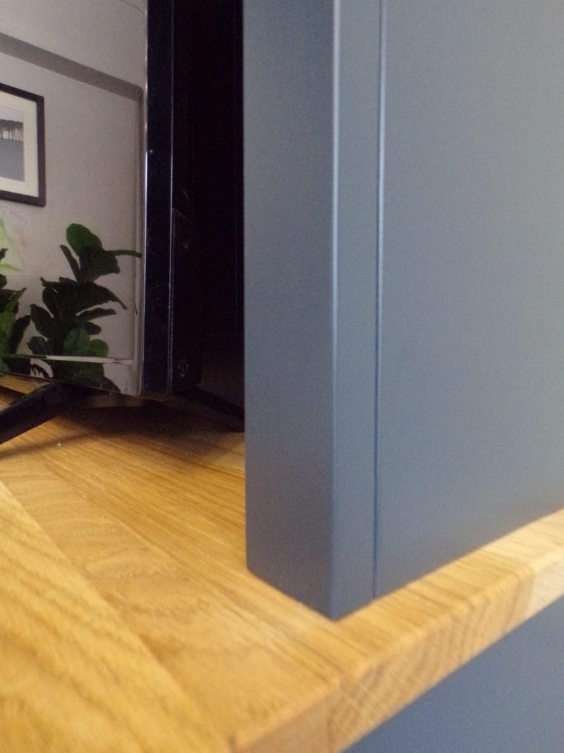Very close view of the oak worktop corner where it meets the navy painted upright, showing the sawn edge and open grain of the oak with the television base and a plant reflected behind