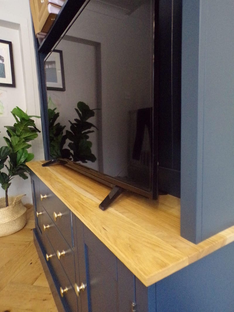 View along the oak worktop from the side, showing the television on its stand in front of the navy backboards, brass knobs on the drawers below and a fiddle leaf fig in a basket beyond