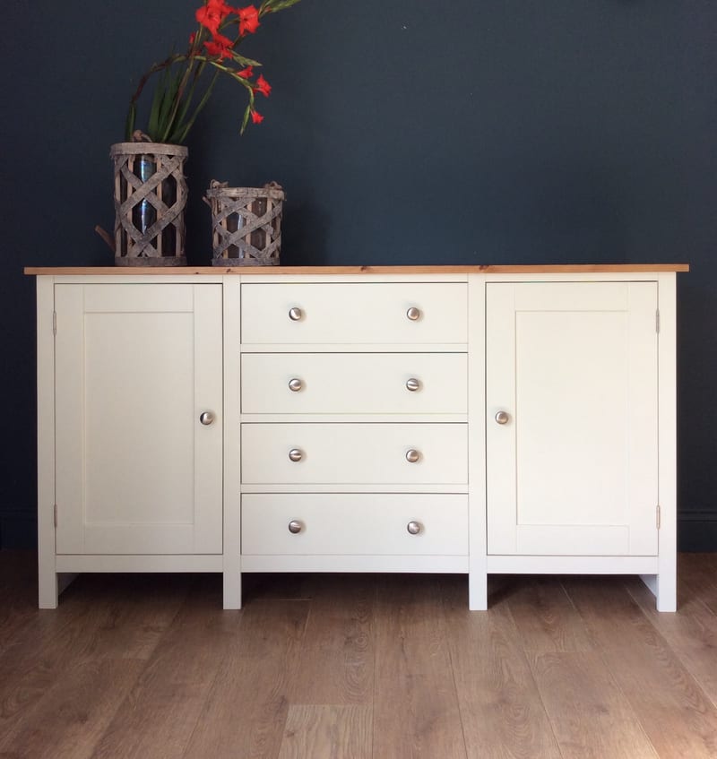 Front view of the cream sideboard on square legs, four drawers between two shaker doors with brushed nickel knobs, and two rustic bark lanterns on the pine top against a dark navy wall