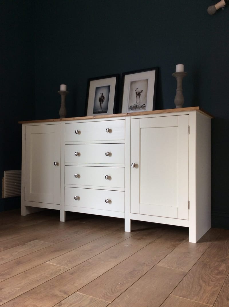 Three-quarter view of the cream sideboard on square legs, showing framed prints and candlesticks on the pine top, four drawers with nickel knobs and two panelled doors on oak flooring