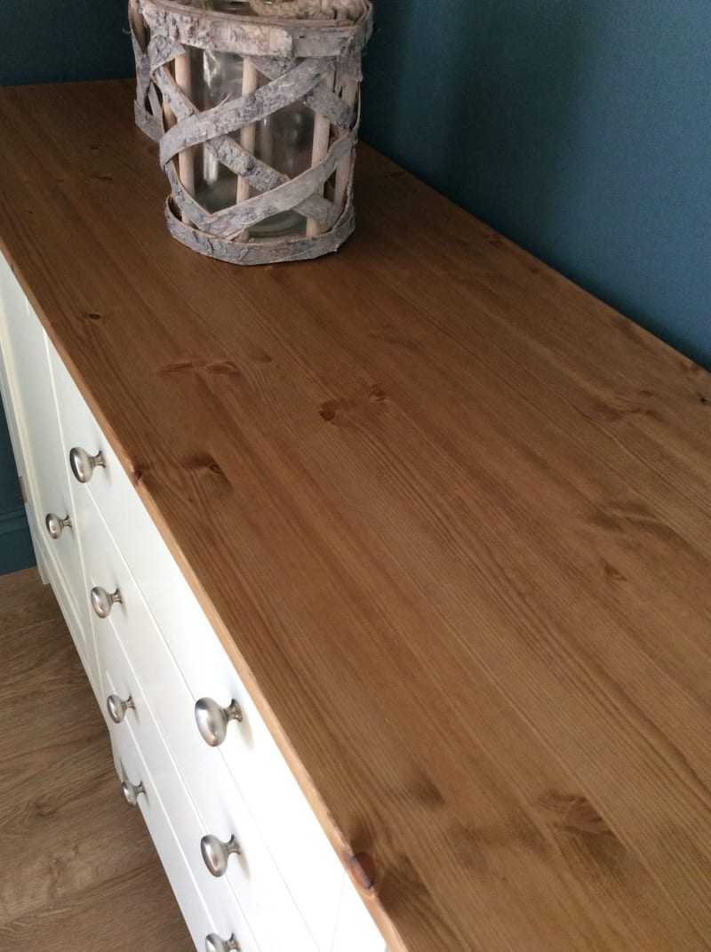Looking along the stained pine top of the cream sideboard, showing the knotty grain and board joins, a rustic bark and glass lantern and the brushed nickel drawer knobs below