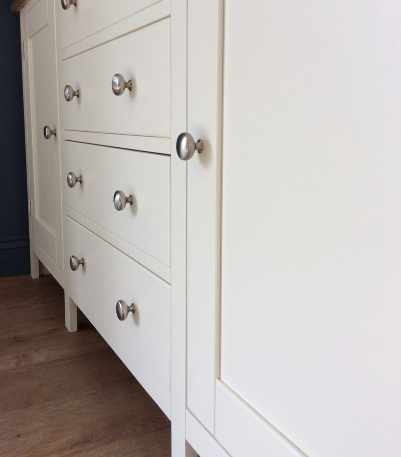 Acute angled view along the cream painted front, showing the run of brushed nickel knobs on the drawers, the flat panelled door beyond, a square leg and the dark navy wall behind