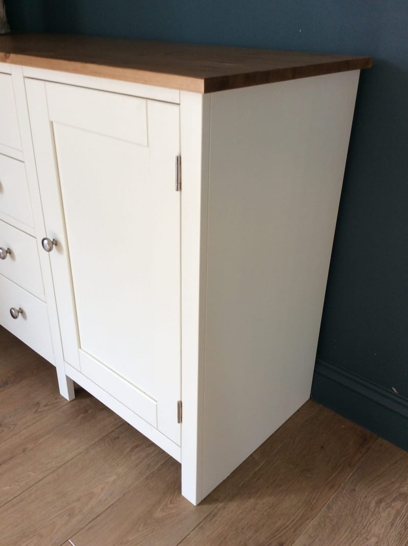 End-on corner view showing the plain painted side of the cream sideboard, the overhanging dark stained pine top, a shaker door on brushed nickel hinges and two square legs on oak flooring