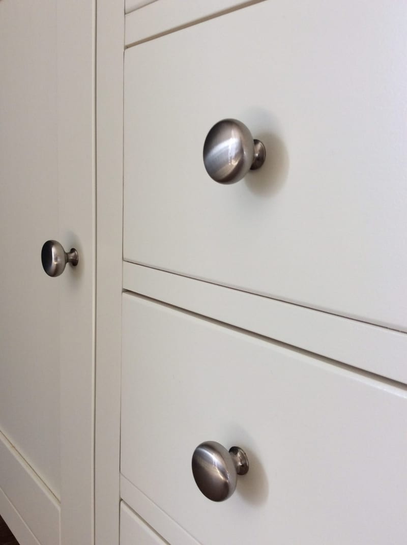 Close-up of two brushed nickel mushroom knobs on the cream drawer fronts next to the framed door stile, showing the fine shadow gaps and the smooth eggshell painted surface