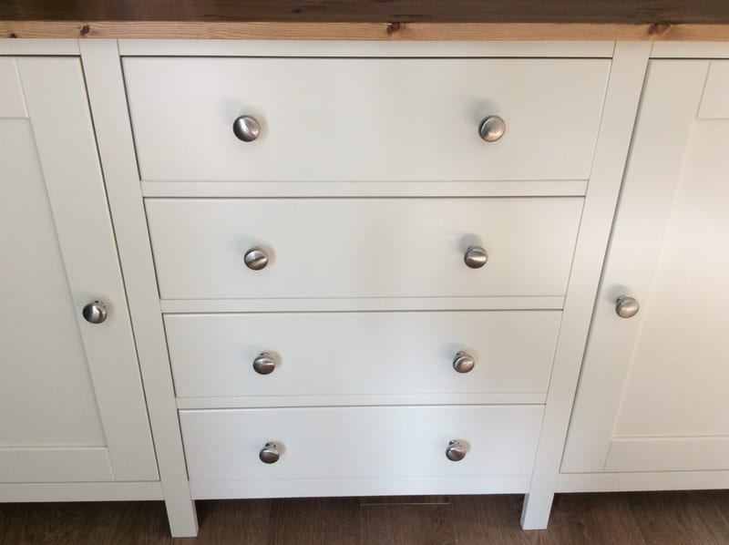 Straight-on close view of the four graduated drawers with pairs of brushed nickel knobs, flanked by the two cupboard doors, with the pine top above and the square legs on dark flooring