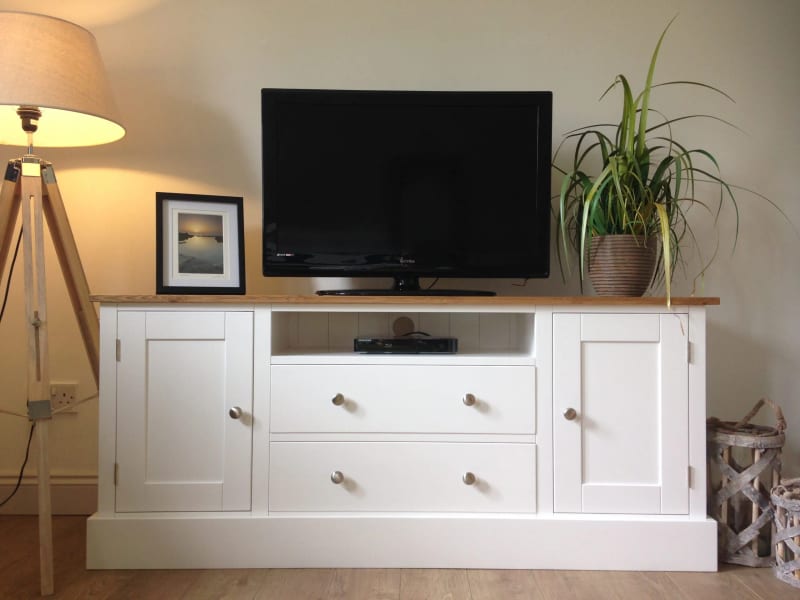 White painted 5ft TV media base unit with an oak top, an open media shelf above two drawers and a cupboard at each end, with a television and a framed print on top
