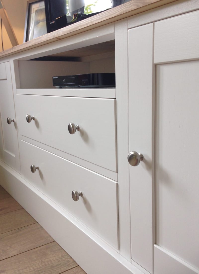 Angled close view along the front of the white painted TV unit, showing two plain drawer fronts with brushed nickel knobs, the open media shelf holding a black Blu-ray player and a shaker side door