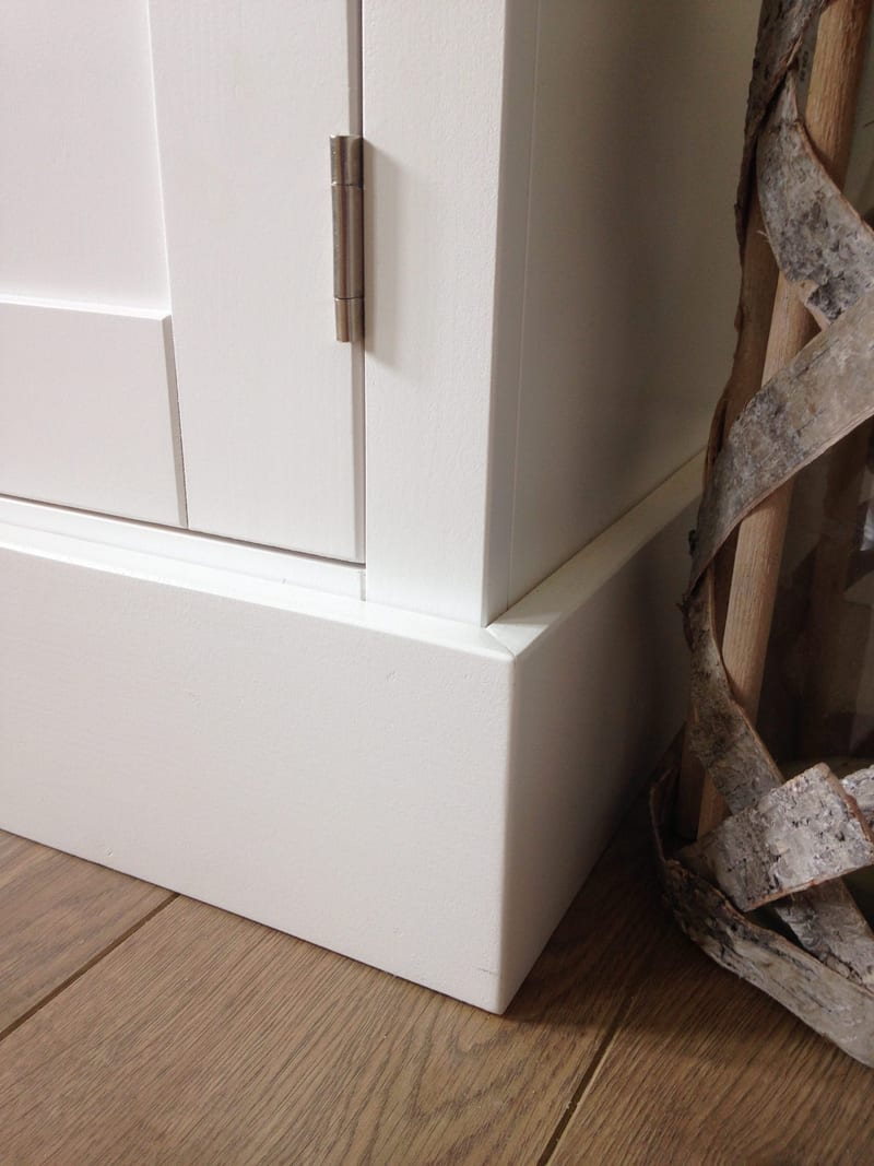 Close view of the bottom corner of the white painted cabinet, showing the plinth, the brushed nickel butt hinge on the shaker door and a woven birch bark basket standing on oak flooring