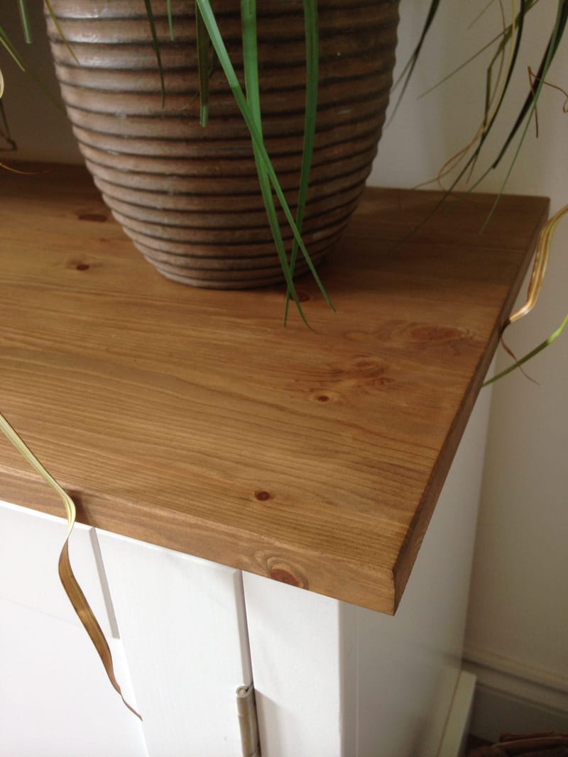 Detail of the corner of the waxed solid pine top, showing its knots, grain and chamfered edge beneath a ribbed terracotta plant pot, with the white painted doors visible below