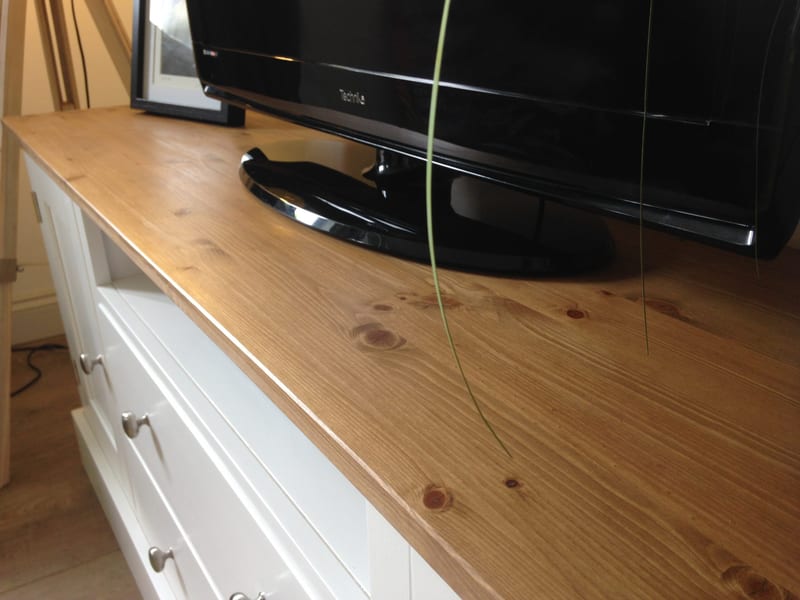 Low angled view across the waxed pine top of the TV unit, with a black flat-screen television on its stand, a framed print behind and the white drawers and nickel knobs at the left