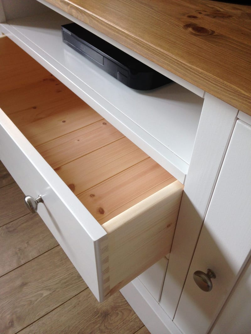 Detail of a drawer pulled open on the white painted TV unit, revealing the natural pine drawer box and hand-cut dovetail joints, with a black Blu-ray player on the shelf above