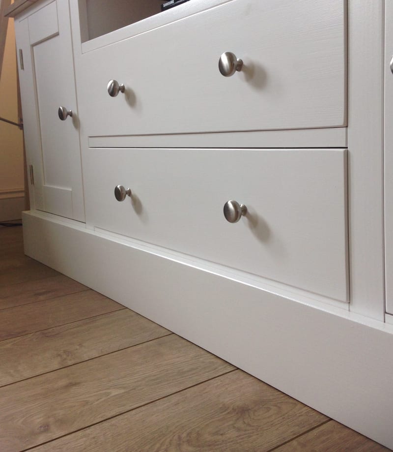 Low view along the two plain drawer fronts of the white painted TV unit, showing the brushed nickel knobs, the shaker cupboard door at the left and the plinth above plank-effect flooring