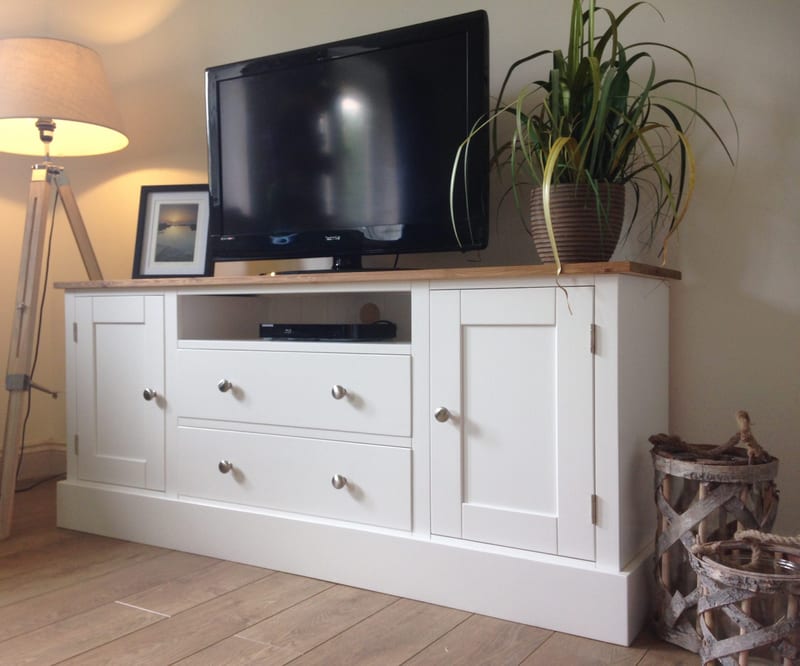 Three-quarter view of the white painted TV unit with waxed pine top, two doors, two drawers and a central media shelf, dressed with a television, a trailing plant and a tripod floor lamp