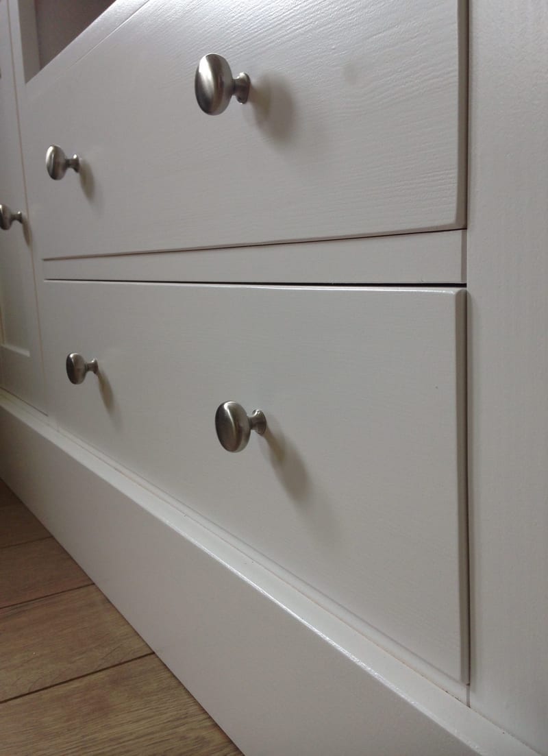 Close-up of two painted drawer fronts on the TV unit, lit from the side to show the brushed nickel knobs, the crisp shadow gaps between drawers and the moulded plinth beneath
