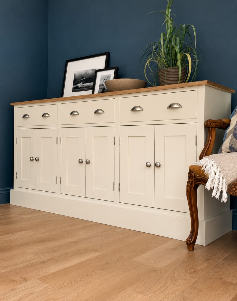 Three-quarter view of the cream painted sideboard from the right, showing the oak top, deep plinth and plain painted end, with a carved armchair and fringed cream throw in the corner