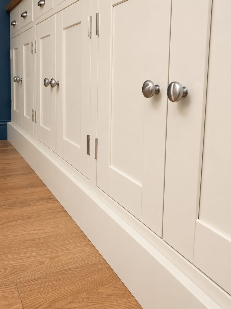 Low raking view along the closed cupboard doors, showing the brushed nickel knobs and butt hinges, the shadow lines of the shaker frames and the plinth meeting the oak flooring